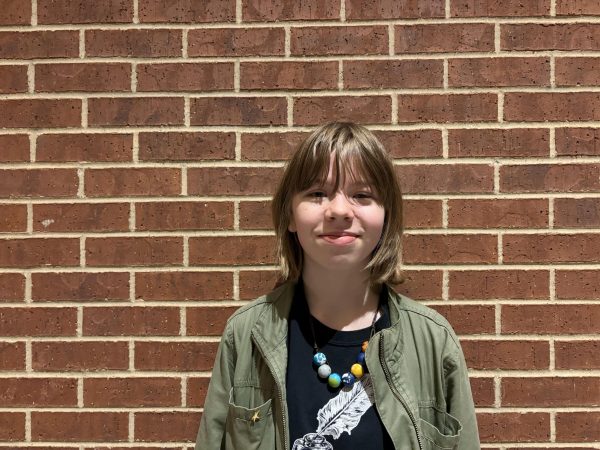 Wearing her creative writing club T-Shirt, junior Macy Washburn stands against a brick wall.